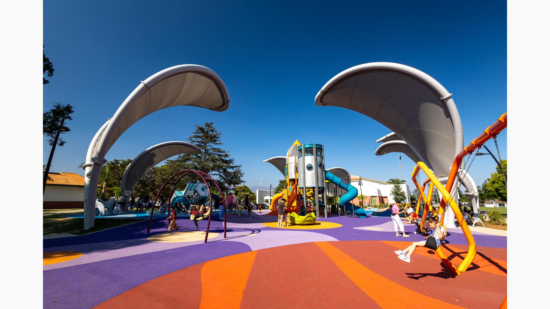 Children play on a colorful playground with large arched shade structures shaped like grey leaves.