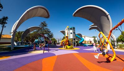 Children play on a colorful playground with large arched shade structures shaped like grey leaves.