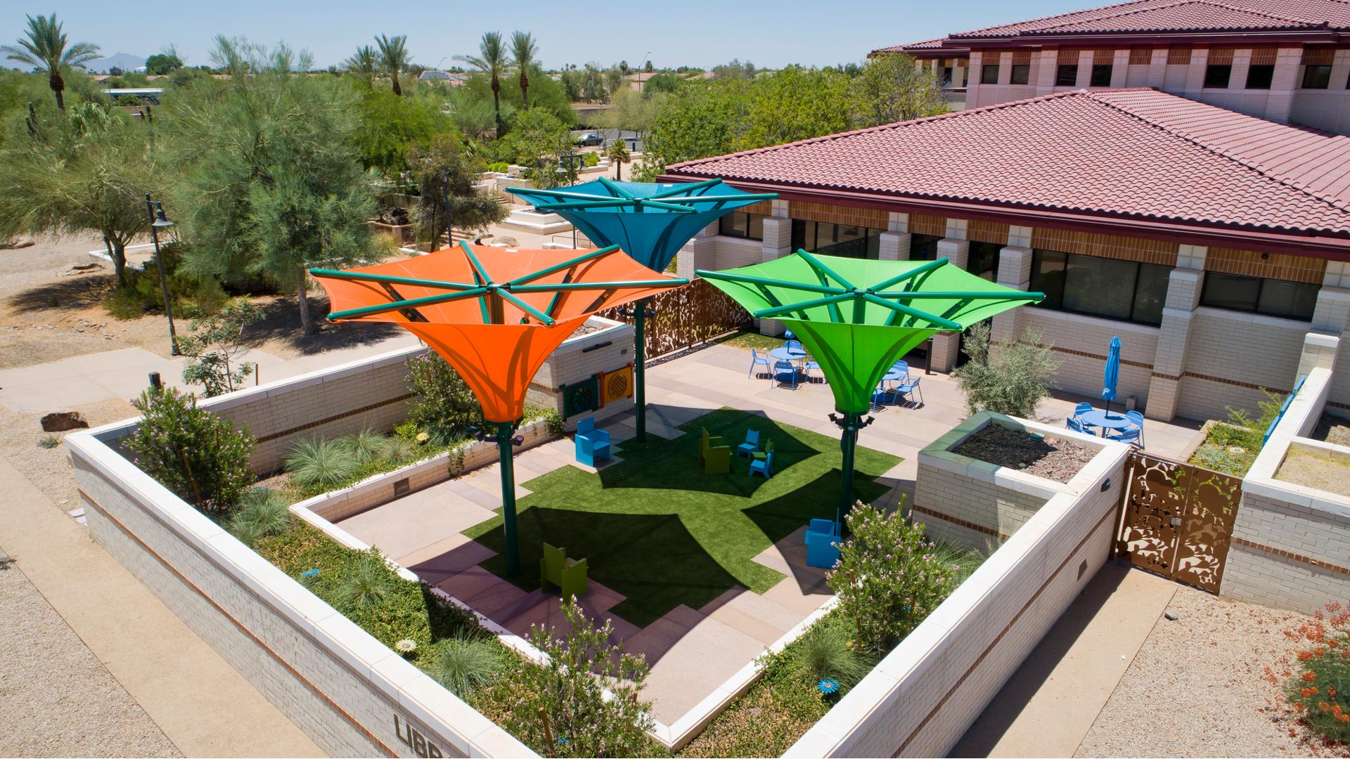 Concrete walls surround a square outdoor garden and sitting area outside of a library with Spanish style tiled roof. Three large colorful shades designed like an inverted umbrella shade the outdoor sitting area.