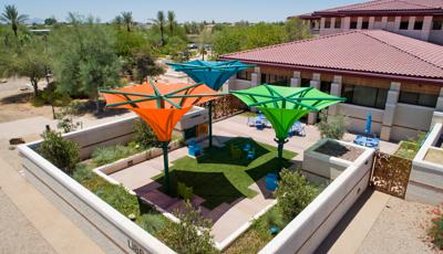 Concrete walls surround a square outdoor garden and sitting area outside of a library with Spanish style tiled roof. Three large colorful shades designed like an inverted umbrella shade the outdoor sitting area.