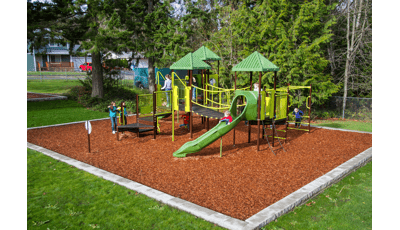 Kids playing on nature inspired playground with green roofs and play equipment. Brown stairs with linking bridge. Tall evergreen trees surround the playground while three girls play chase across the wood chips around the playground.