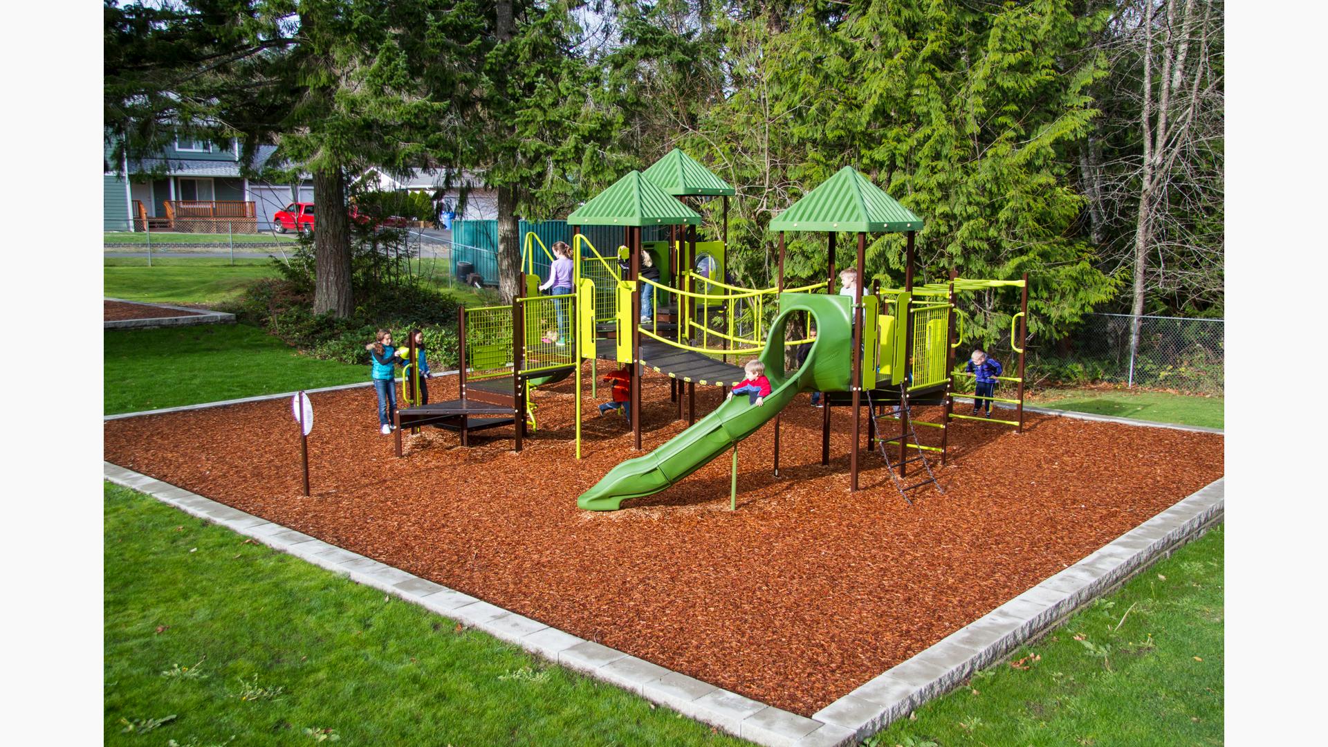 Kids playing on nature inspired playground with green roofs and play equipment. Brown stairs with linking bridge. Tall evergreen trees surround the playground while three girls play chase across the wood chips around the playground.