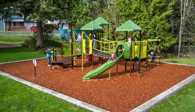 Kids playing on nature inspired playground with green roofs and play equipment. Brown stairs with linking bridge. Tall evergreen trees surround the playground while three girls play chase across the wood chips around the playground.