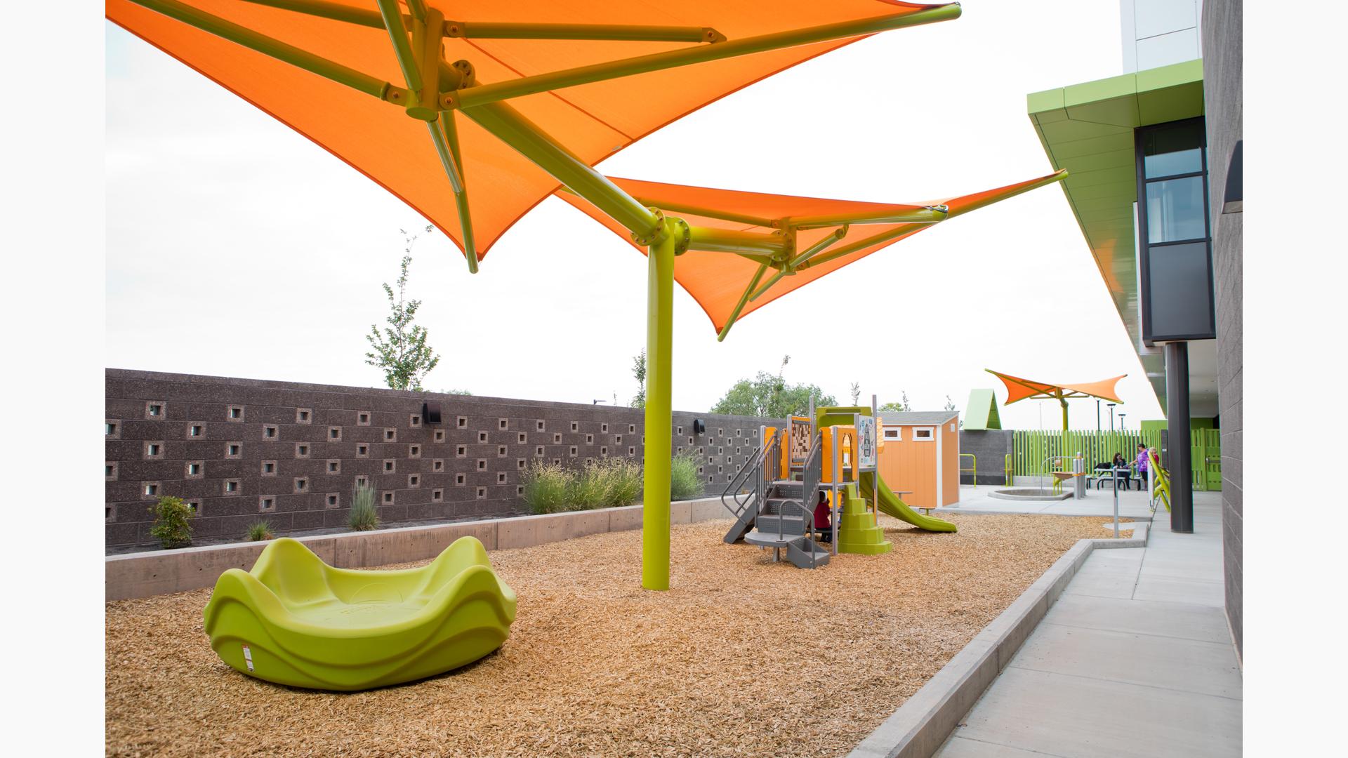 A green multi seat spinner sits under a brightly colored shade system with a small play structure in the background. The play area sits between a granite retaining wall and a building.
