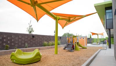 A green multi seat spinner sits under a brightly colored shade system with a small play structure in the background. The play area sits between a granite retaining wall and a building.