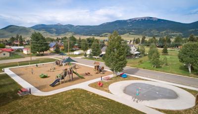 Elevated view of a play area with play structures and other freestanding play activities next to a splash pad with ground water sprayers. Neighborhood homes fill the background leading to a mountainous hillside.