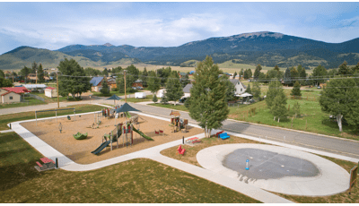 Elevated view of a play area with play structures and other freestanding play activities next to a splash pad with ground water sprayers. Neighborhood homes fill the background leading to a mountainous hillside.