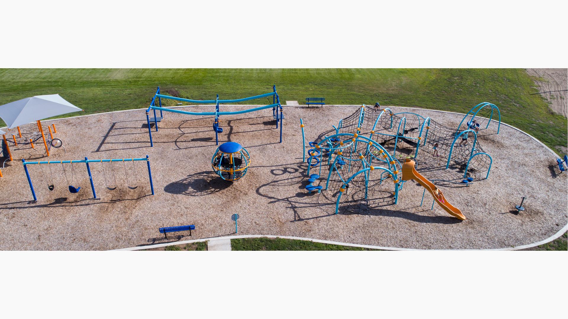 Elevated view of a park playground with blue posts as children climb through the weaving ropes and belt climbers of the play structure.