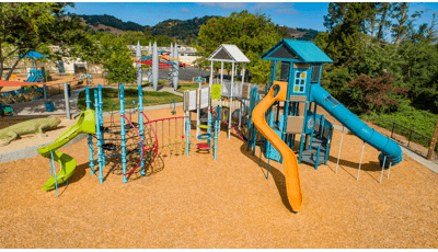 Two colorful tower structures connected by bridges and climbers great a large playground all on a wood chip surfacing. 
