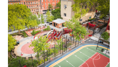 Aerial view of a playground park amongst city buildings surrounded by black wire fencing next to multiple basketball courts.
