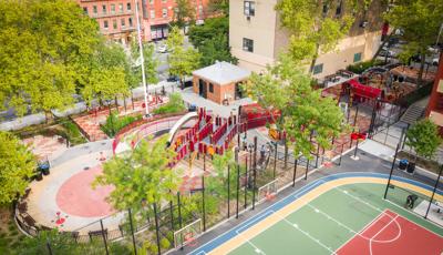 Aerial view of a playground park amongst city buildings surrounded by black wire fencing next to multiple basketball courts.