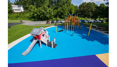 Orange dynamic rope structure is nested in neighborhood park with red playhouse for younger kids all with blue poured-in-place playground surface. 