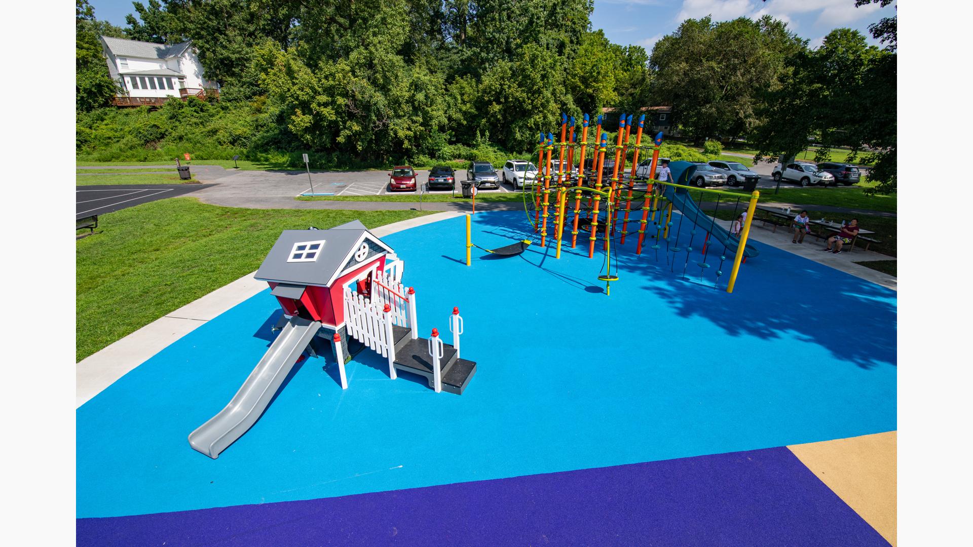 Orange dynamic rope structure is nested in neighborhood park with red playhouse for younger kids all with blue poured-in-place playground surface. 
