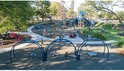 A large park surrounded by mature trees that shade the entire play area. Families sit at picnic tables and chairs around and scattered amongst the play area. Children play on a swing sets made up of three arch bays while a crowd of people play on the main large play structure with a tower.