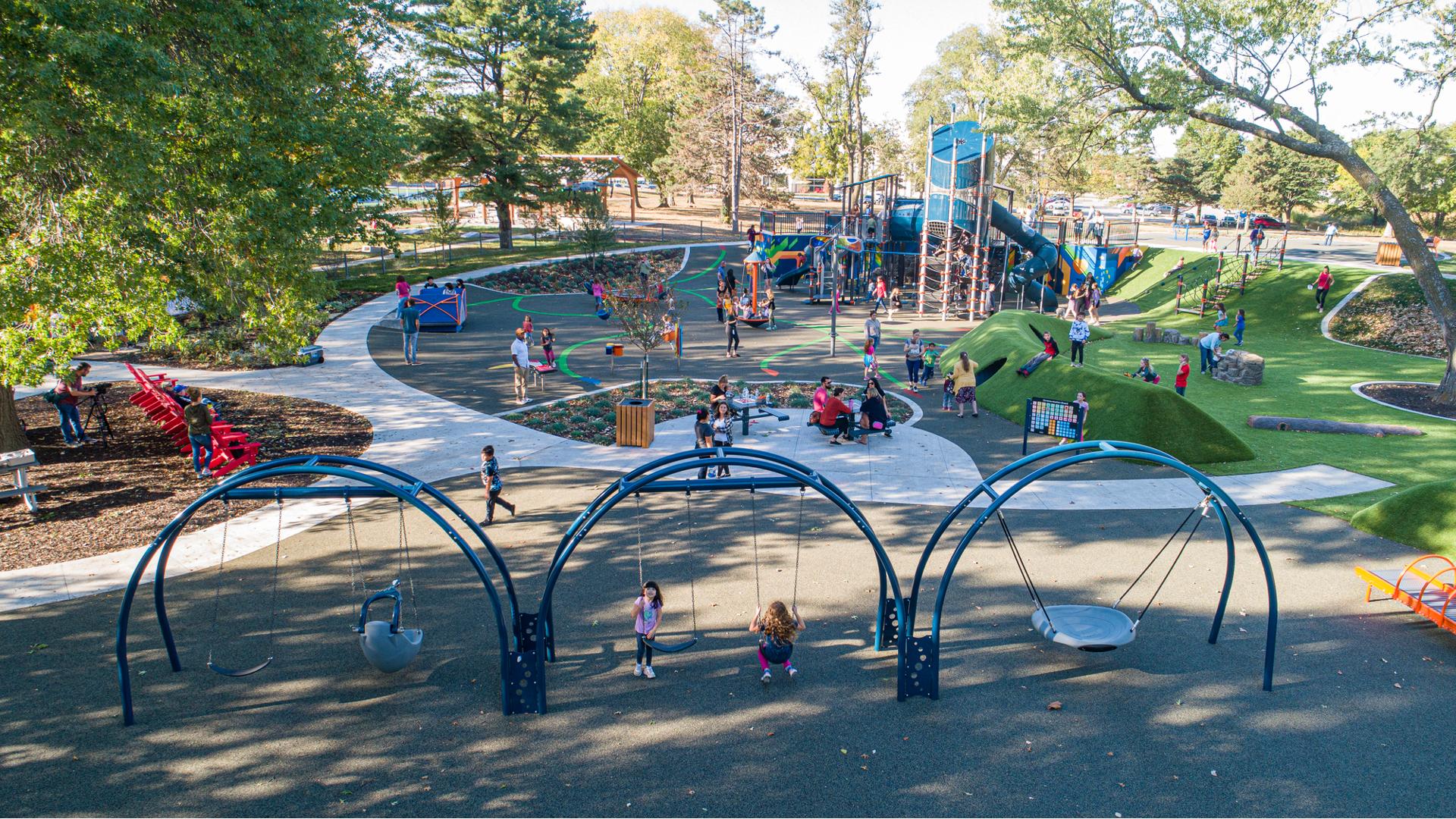 A large park surrounded by mature trees that shade the entire play area. Families sit at picnic tables and chairs around and scattered amongst the play area. Children play on a swing sets made up of three arch bays while a crowd of people play on the main large play structure with a tower.