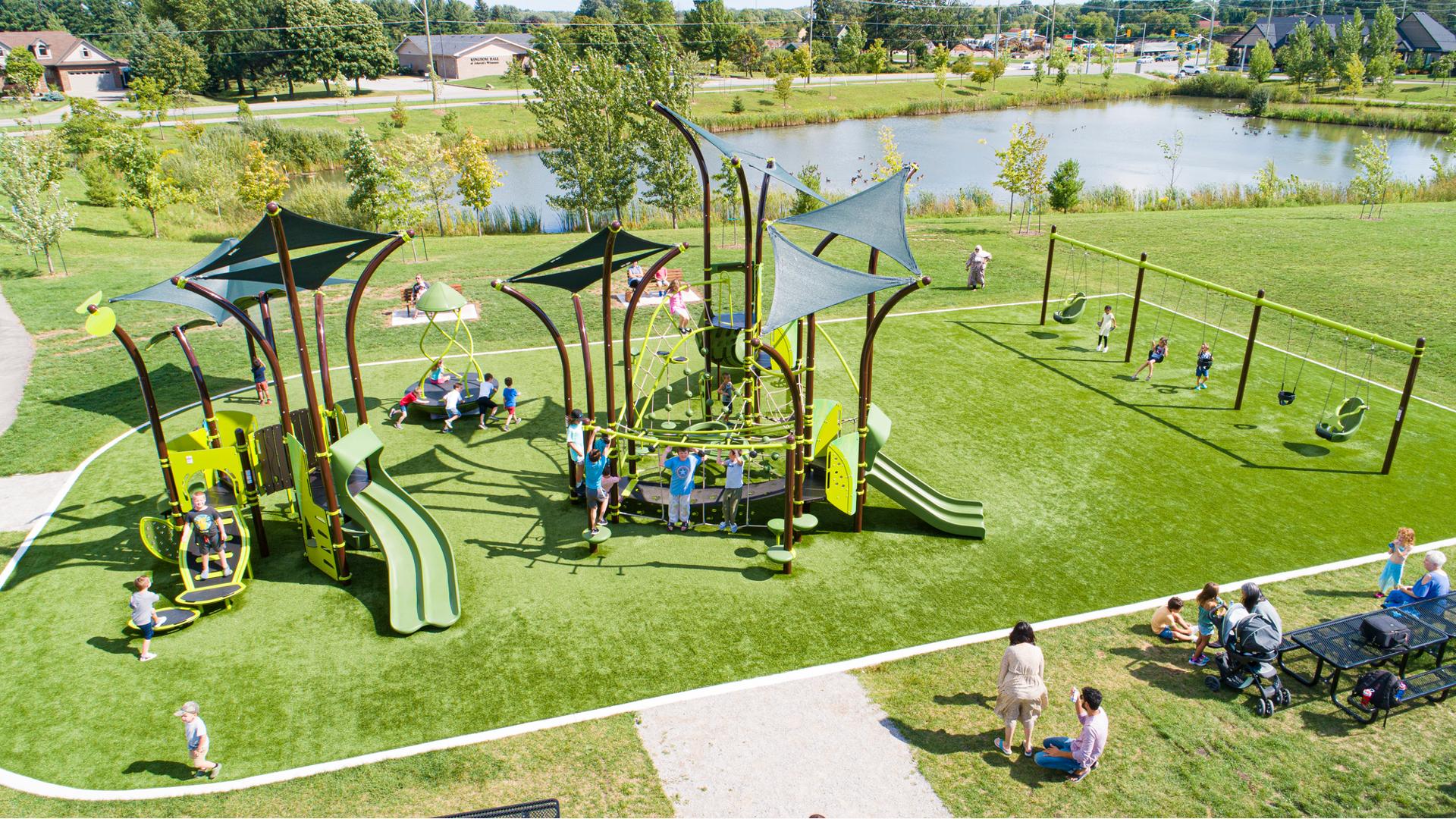 Families sit at picnic tables on the side of a park play area with nature inspired play structures, row of swings sets and a large pond in the background. 