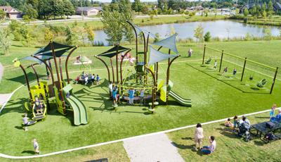 Families sit at picnic tables on the side of a park play area with nature inspired play structures, row of swings sets and a large pond in the background. 