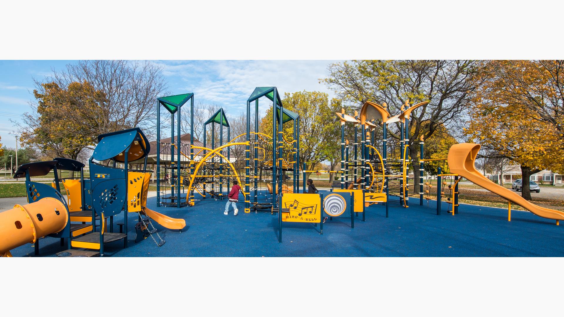 Children run on the blue playground safety surfacing amongst the navy blue and gold colored play structures with rope climbers, play panels, and slides.