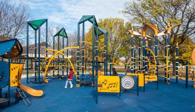 Children run on the blue playground safety surfacing amongst the navy blue and gold colored play structures with rope climbers, play panels, and slides.