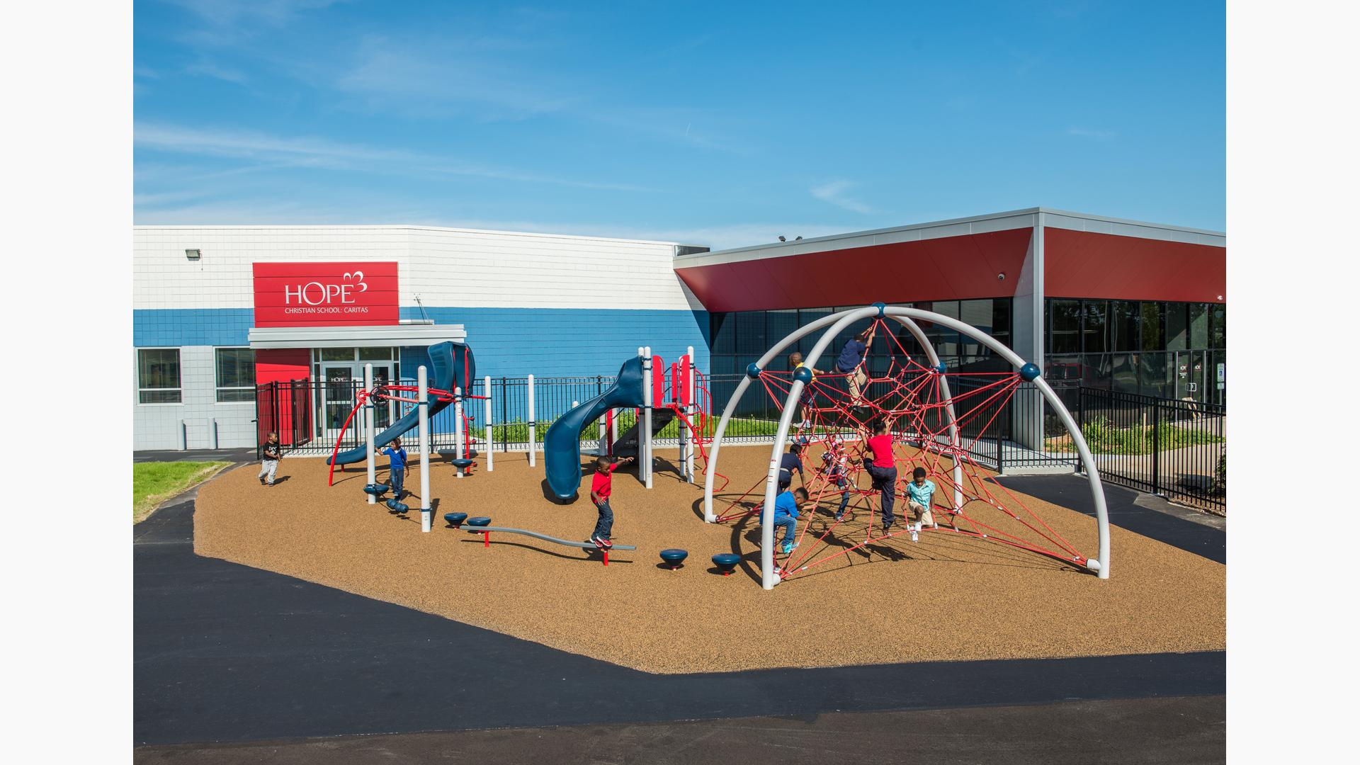 Children play on bridges, steppers, slides, and balance beams all leading to a large net climber next to a school building. 