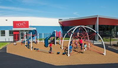Children play on bridges, steppers, slides, and balance beams all leading to a large net climber next to a school building. 