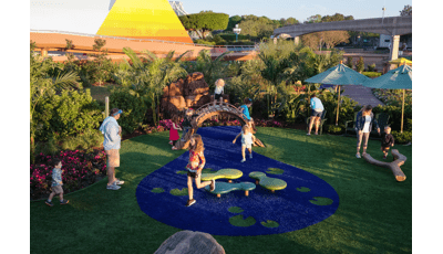 Parents watch as children play on nature themed wobblers and log balance beams on artificial grass designed as a pond of water with lily pads all surrounded by a tropical garden.