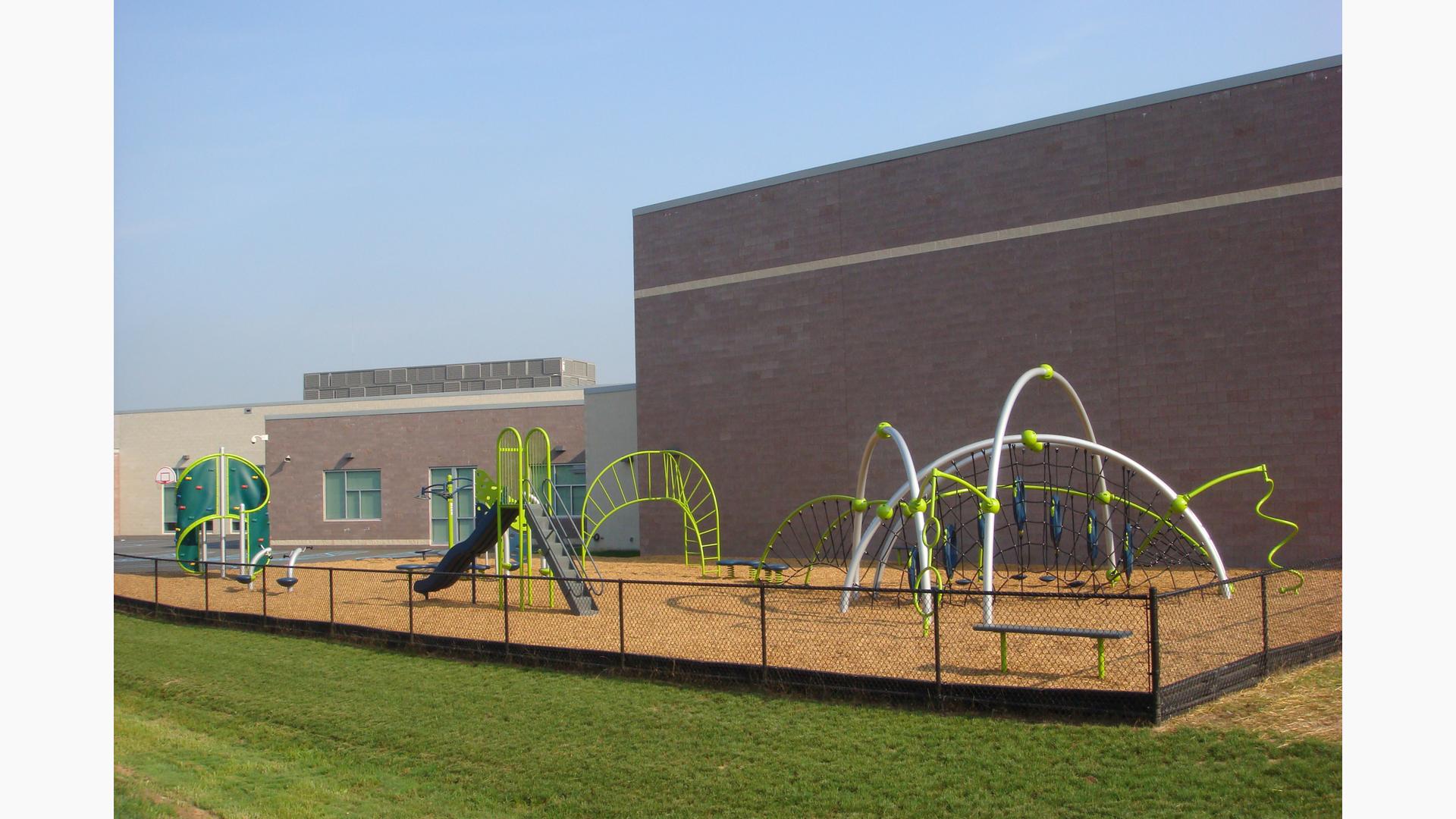 The playground rests behind the school. several Evos and PlayBooster structures sit fenced in with a bright green lawn just beyond the fencing.