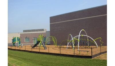 The playground rests behind the school. several Evos and PlayBooster structures sit fenced in with a bright green lawn just beyond the fencing.