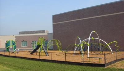 The playground rests behind the school. several Evos and PlayBooster structures sit fenced in with a bright green lawn just beyond the fencing.
