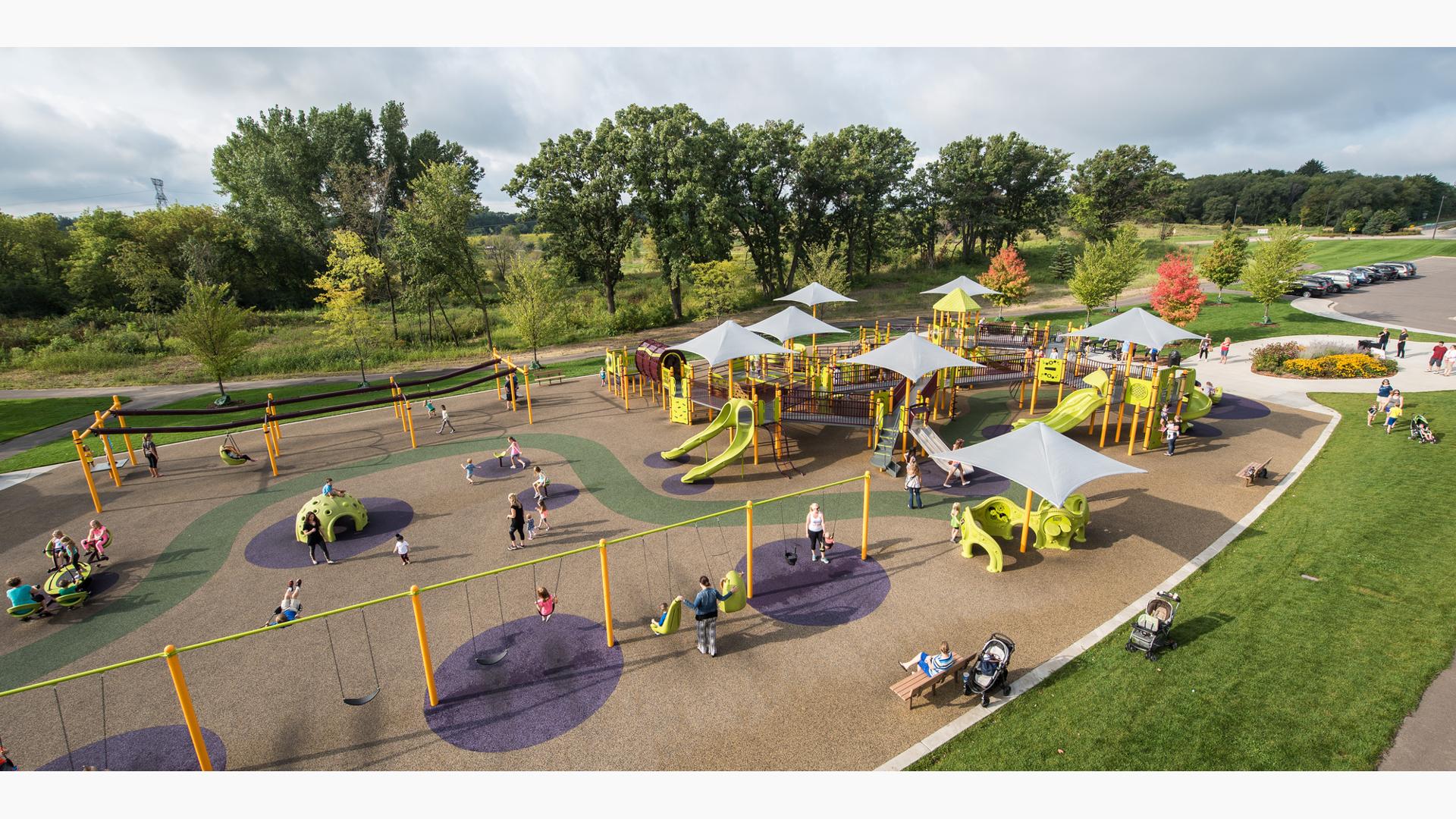 Aerial view of large playground with kids and families playing on swings, ziplines and playground. Playground is colors include bright green slides and climbers with gray shade. 