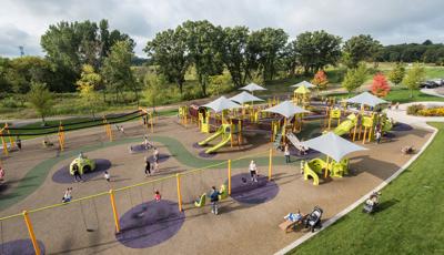 Aerial view of large playground with kids and families playing on swings, ziplines and playground. Playground is colors include bright green slides and climbers with gray shade. 