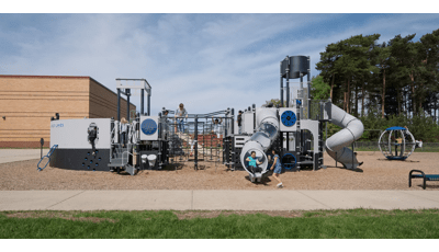 A girl exits tunnel slide from custom Navy ship design PlayBooster play structure at Great Lakes Elementary School. The playground sits on a mulch ground. The school building sits behind the playground as tall trees blow in the wind.