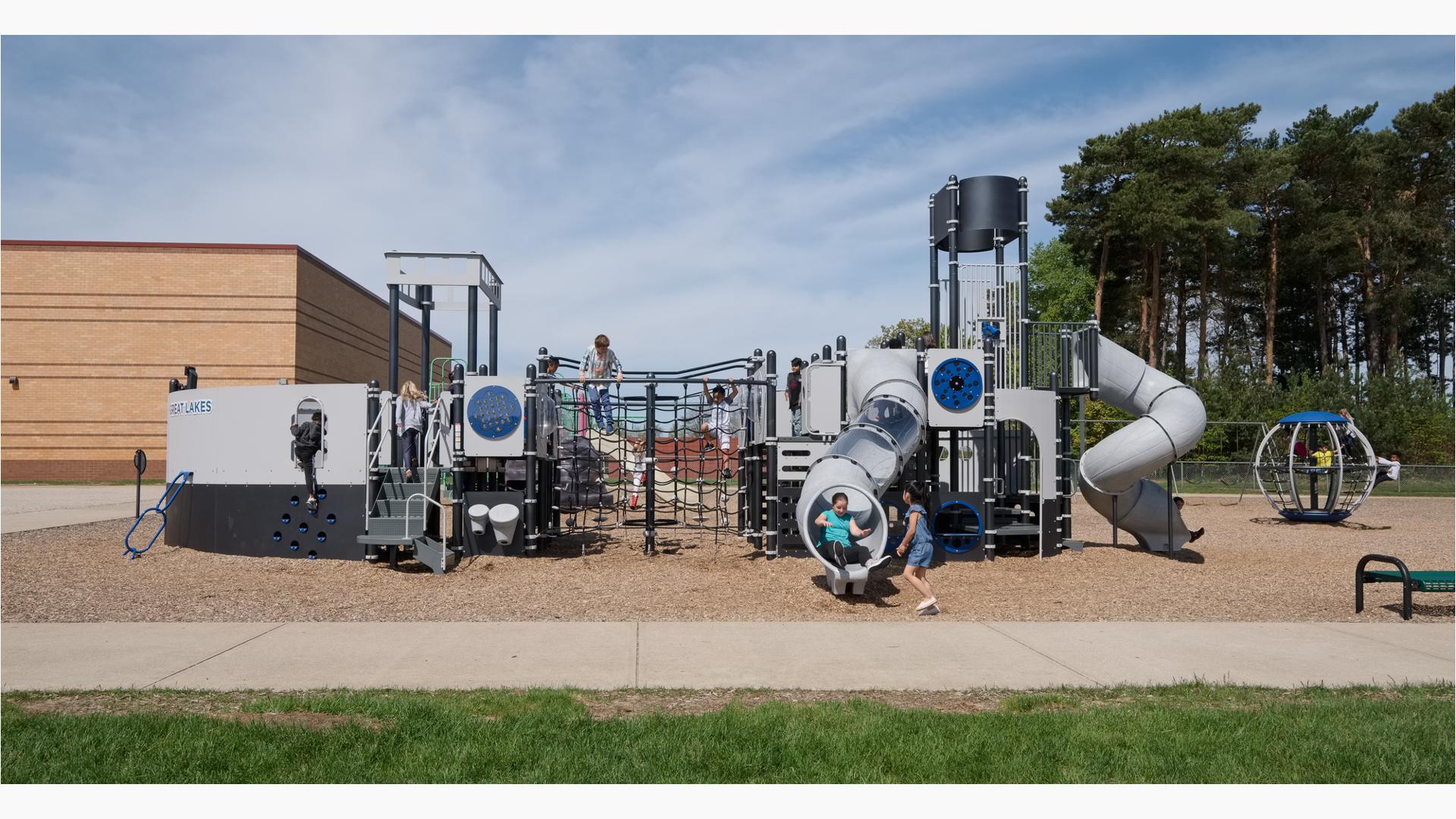 A girl exits tunnel slide from custom Navy ship design PlayBooster play structure at Great Lakes Elementary School. The playground sits on a mulch ground. The school building sits behind the playground as tall trees blow in the wind.