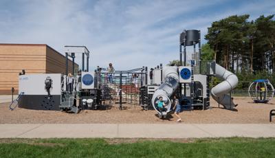 A girl exits tunnel slide from custom Navy ship design PlayBooster play structure at Great Lakes Elementary School. The playground sits on a mulch ground. The school building sits behind the playground as tall trees blow in the wind.