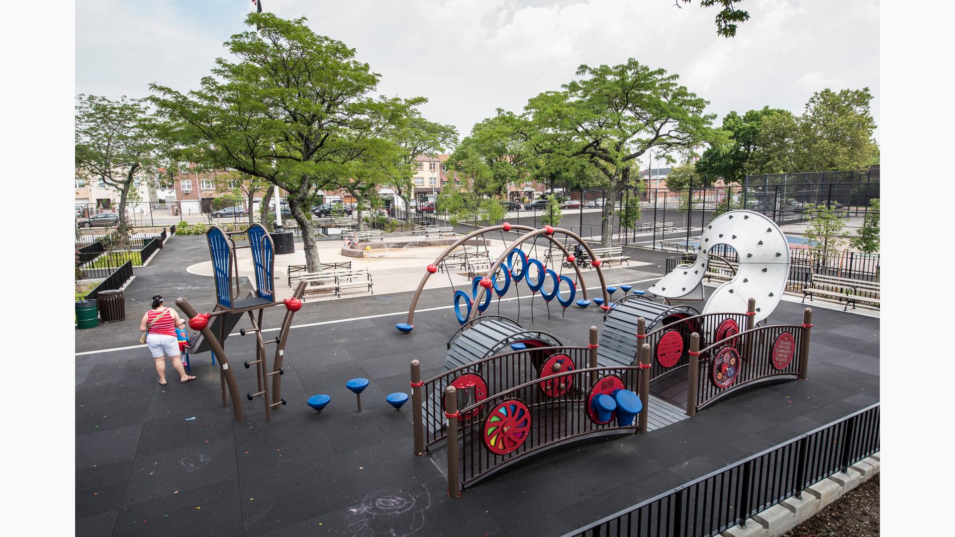 GIrl with her mother stands at foot of slide in urban playground. Paul Raimonda Playground includes a brown bridge with red and blue interactive play panels, a wavy climber, slide, large metal climber with hand and foot holds. 