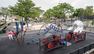 GIrl with her mother stands at foot of slide in urban playground. Paul Raimonda Playground includes a brown bridge with red and blue interactive play panels, a wavy climber, slide, large metal climber with hand and foot holds. 