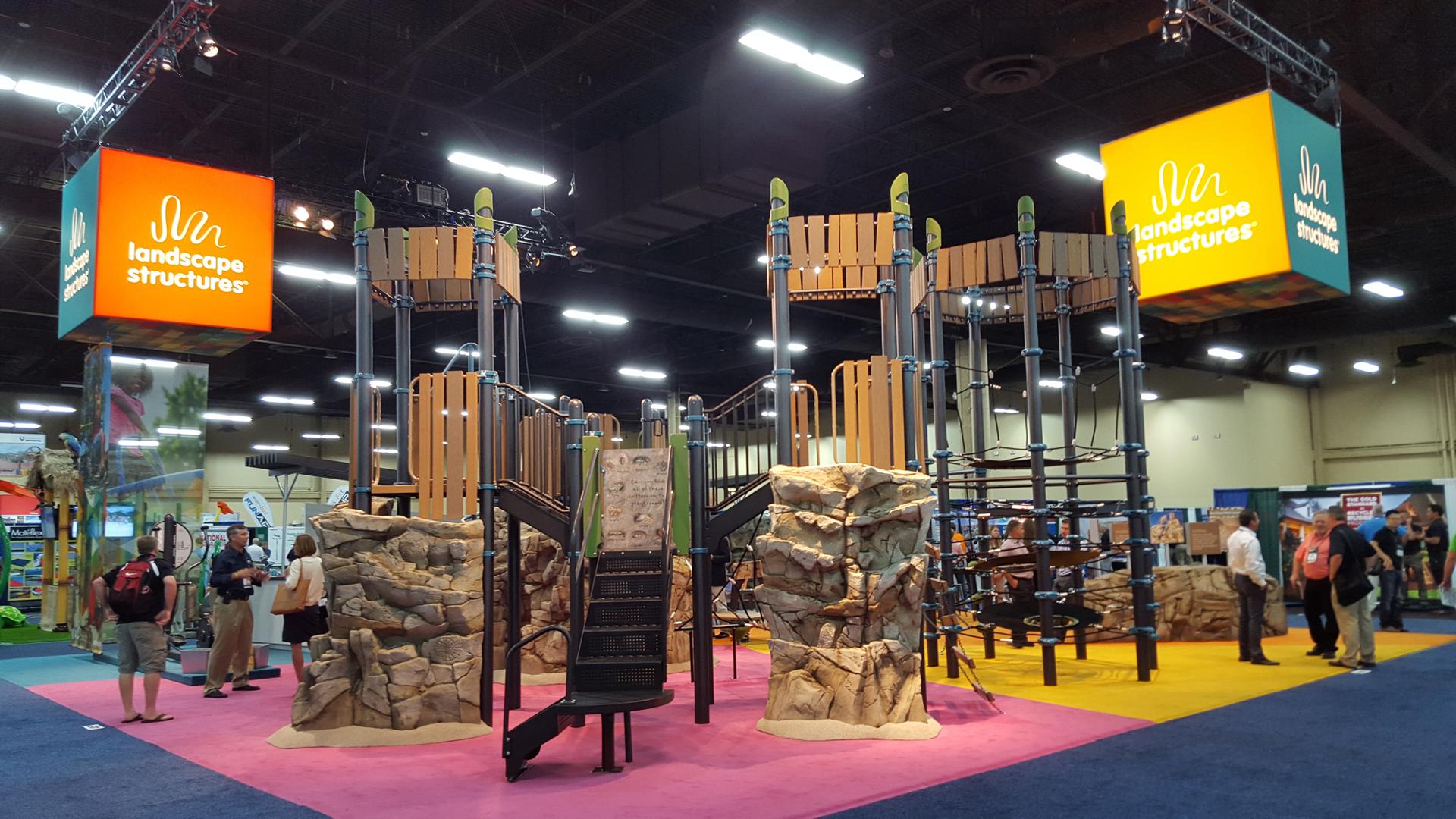 Nature-inspired playground design with real-life looking climbing rocks and recycled wood-grain panels and roofs. Playground is displayed inside a convention center. 