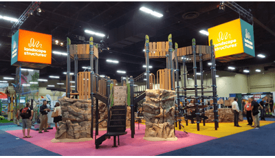 Nature-inspired playground design with real-life looking climbing rocks and recycled wood-grain panels and roofs. Playground is displayed inside a convention center. 