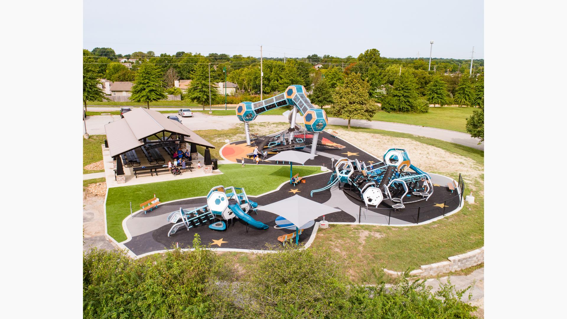 Three geometric blue and silver playground areas for all ages in park location. The playground tower includes three pods with an enclosed connected walkway.  The surfacing is design with stars and planets to round out the space theme play space. 