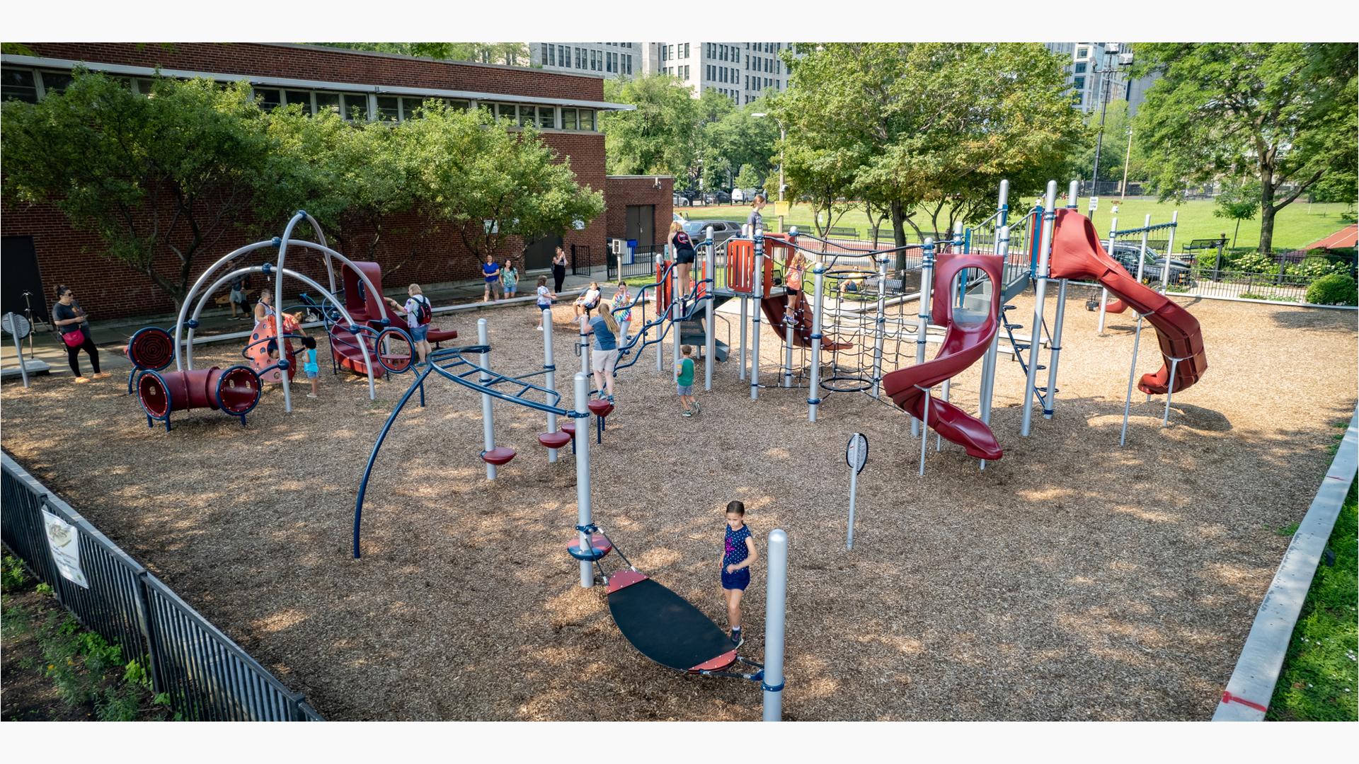 Children play on the many activities of a park playground next to a brick building. 