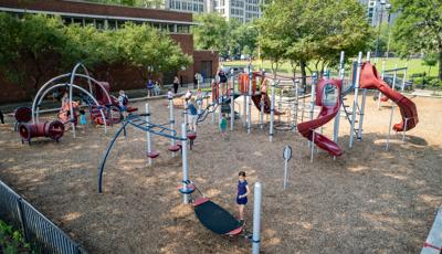 Children play on the many activities of a park playground next to a brick building. 