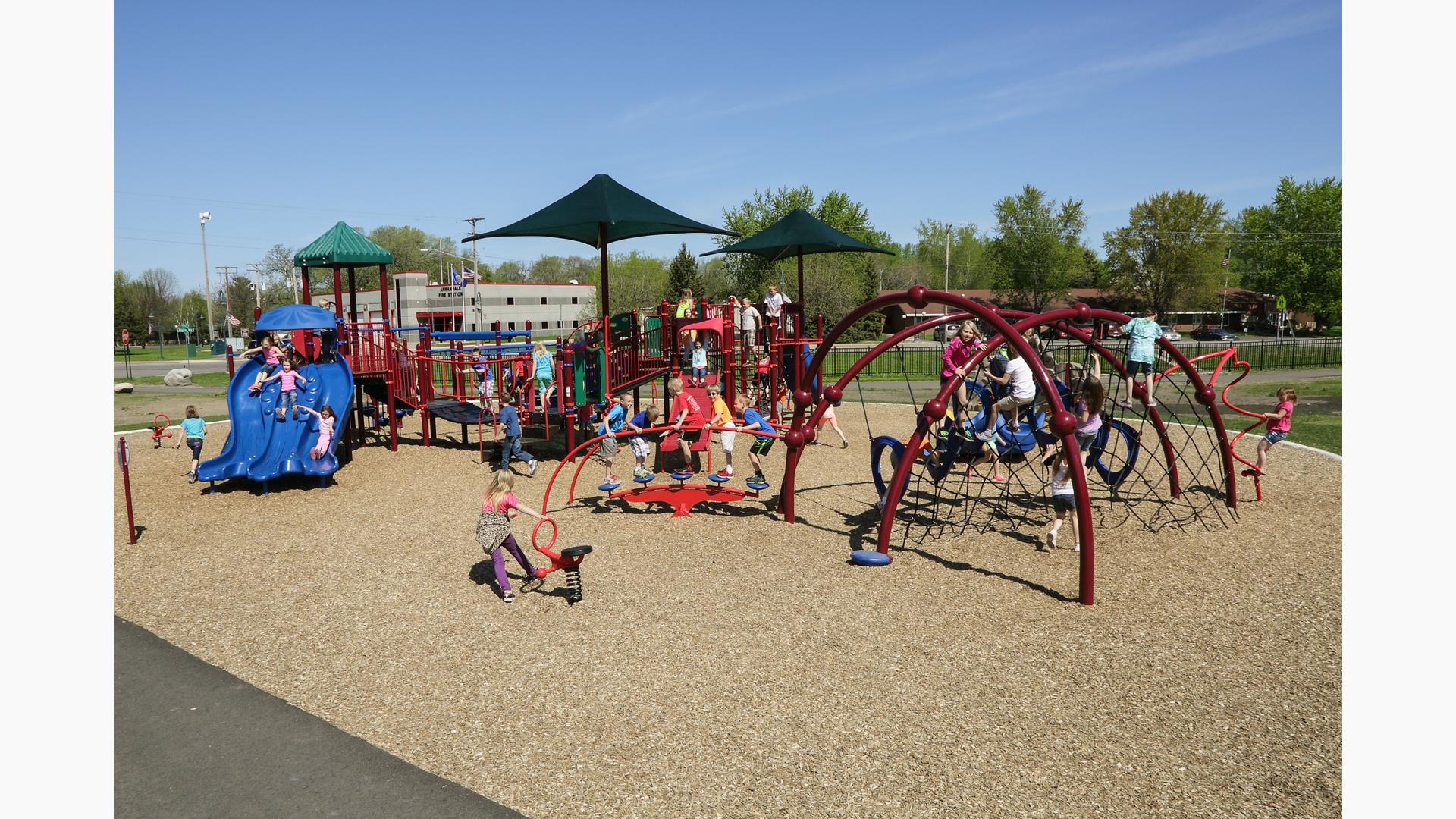 A crowd of children play on a schools two maroon play structures filled with rope climbers, slides, and bridges.