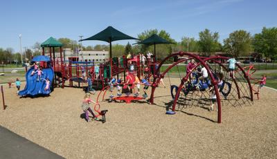 A crowd of children play on a schools two maroon play structures filled with rope climbers, slides, and bridges.