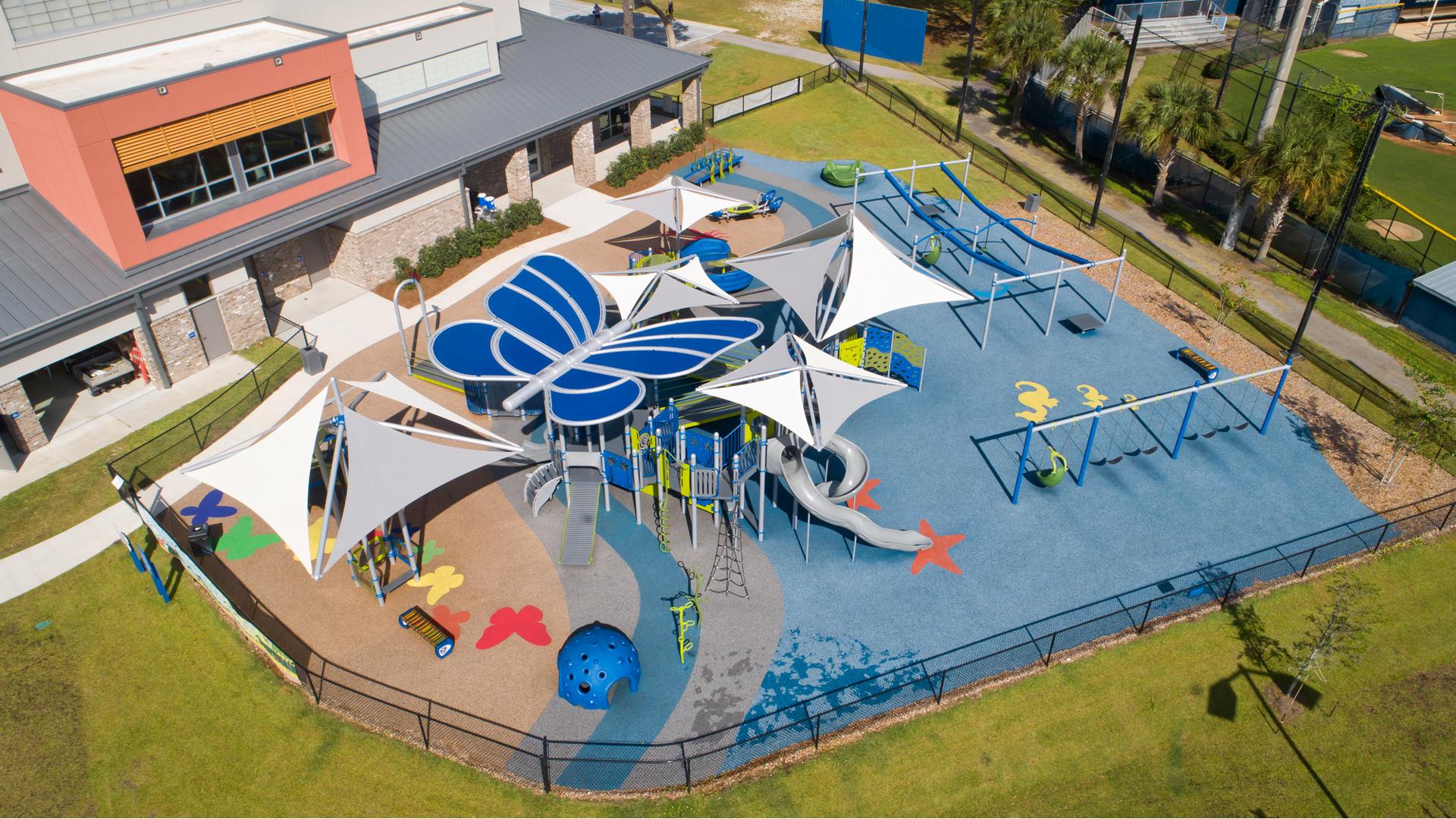 Aerial view of playground with a blue butterfly shaped shade structure. Playground surfacing includes colorful butterflies and starfish all next to school building. 