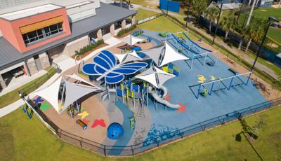 Aerial view of playground with a blue butterfly shaped shade structure. Playground surfacing includes colorful butterflies and starfish all next to school building. 