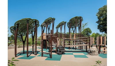Custom tree structures tower over PlayBooster play structure. Log Steppers and rock climbers offer a realistic, natural look to the playground. Play area sits on a green and tan checkered ground surface.