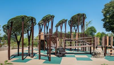 Custom tree structures tower over PlayBooster play structure. Log Steppers and rock climbers offer a realistic, natural look to the playground. Play area sits on a green and tan checkered ground surface.