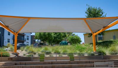 A large rectangular shade protects the concrete seating area of an amphitheater set into a hillside filled with grassy bushes in a neighborhood setting. 