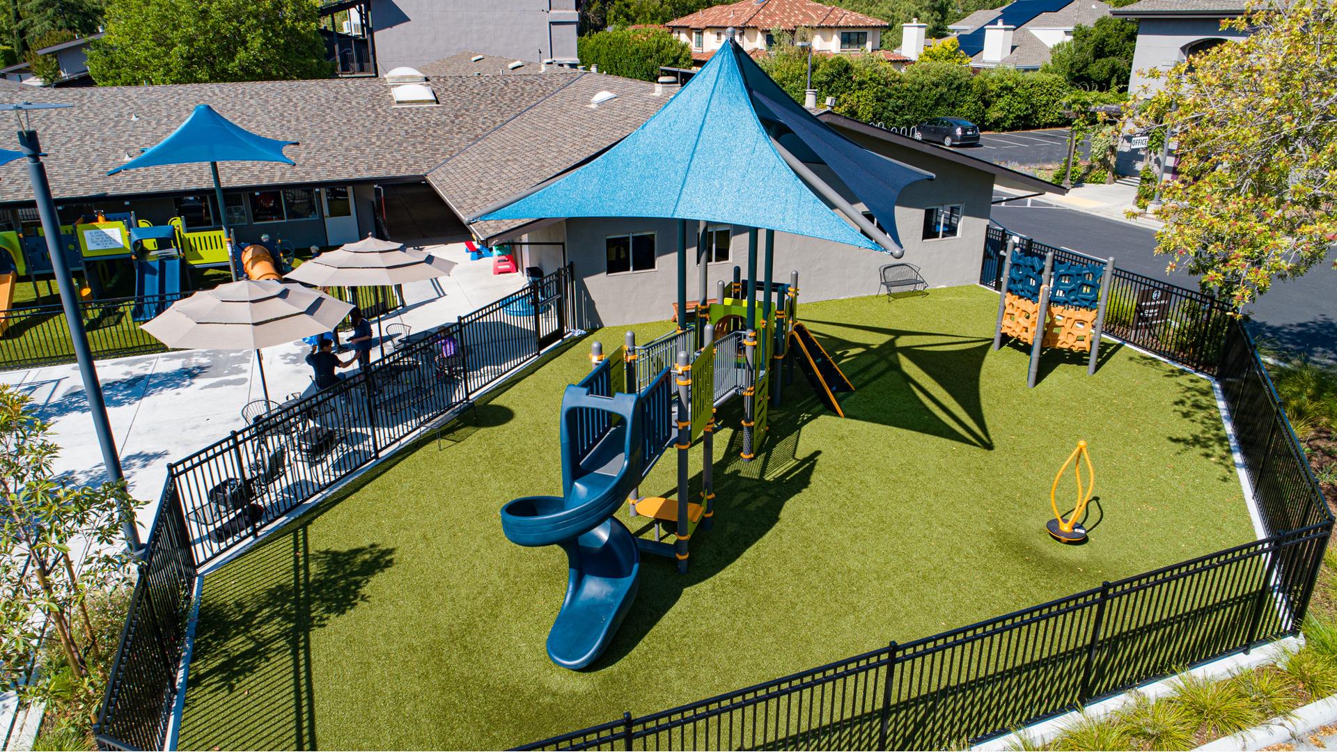Elevated view of a fenced in play area with play structure and large blue overhead shade incorporated next to a church building with a secondary fenced off play area for younger children.