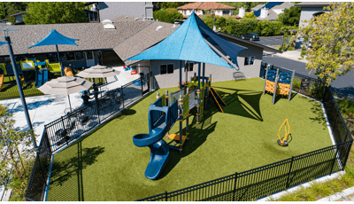 Elevated view of a fenced in play area with play structure and large blue overhead shade incorporated next to a church building with a secondary fenced off play area for younger children.